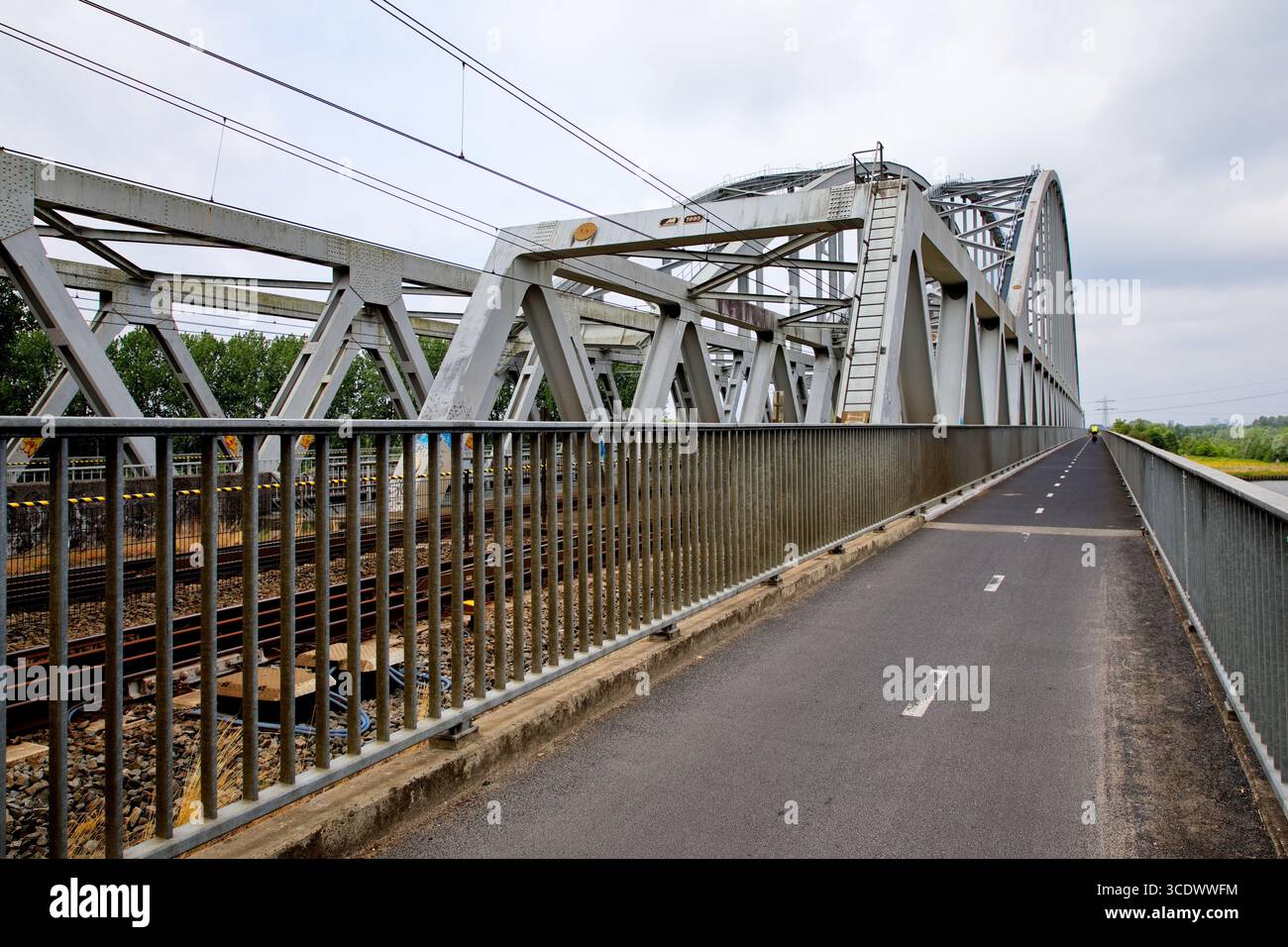 Stahlfachwerkbahnbrücke mit separatem Radweg über den Fluss in den Niederlanden, Konvergenzstrecken und Freileitungen bilden eine Verkehrsszene. Stockfoto