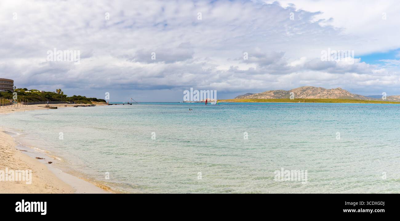 Panoramablick auf den Strand La Pelosa auf Sardinien, mit türkisfarbenem, flachem Wasser, Sandstrand und fernen felsigen Hügeln unter bewölktem Himmel. Stockfoto