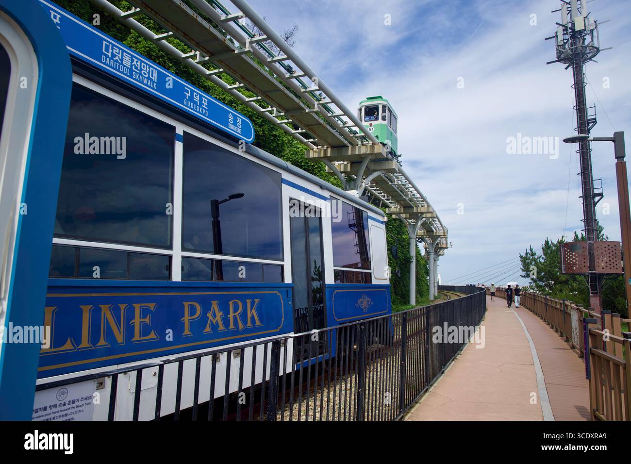 Ein Haeundae Beach Train wartet neben der Strandpromenade, während ein grüner Himmelskapselwagen auf dem erhöhten Gleis in der Nähe des Daritdol Skyw fährt Stockfoto