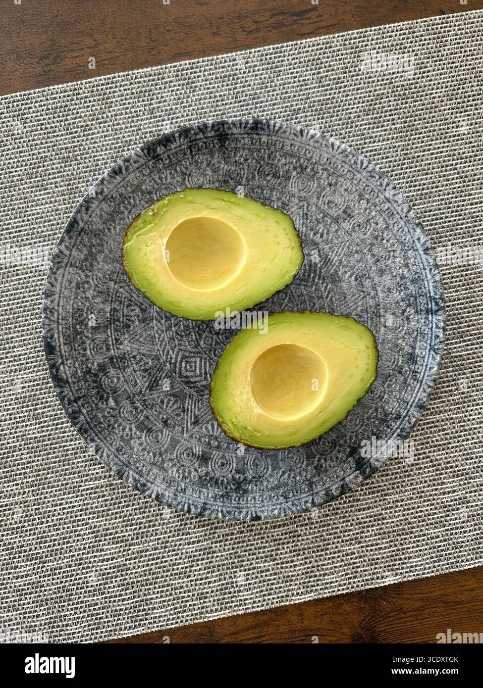 Reife Avocado in zwei Hälften geschnitten, auf einem dekorativen grauen Teller über einem strukturierten Tischset auf einem Holztisch platziert, gesunde Bio-Lebensmittel, Blick von oben - Smartphone-aufgenommenes Stockfoto