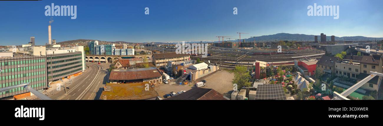 Weitwinkelpanorama mit einer urbanen Szene mit Bahngleisen und markanter Architektur, eingerahmt unter einem leuchtend blauen Himmel. Stockfoto