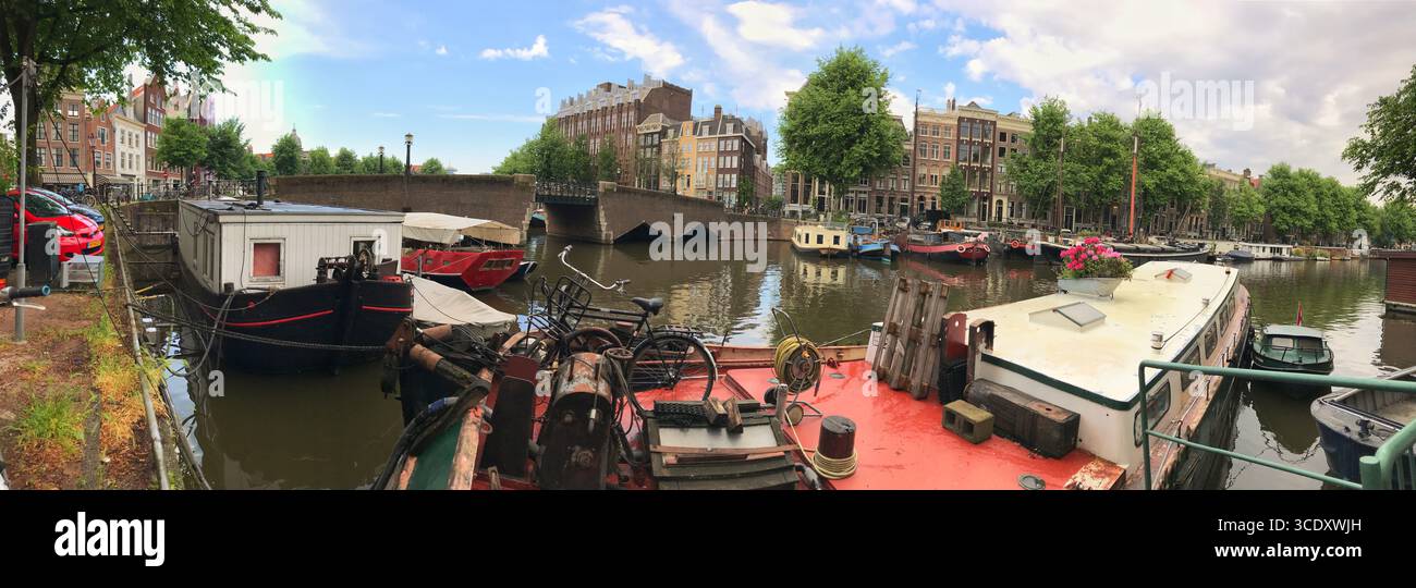 Weite Aussicht auf einen friedlichen Kanal, flankiert von historischen Fassaden und Hausbooten unter einem teilweise bewölkten Himmel. Stockfoto