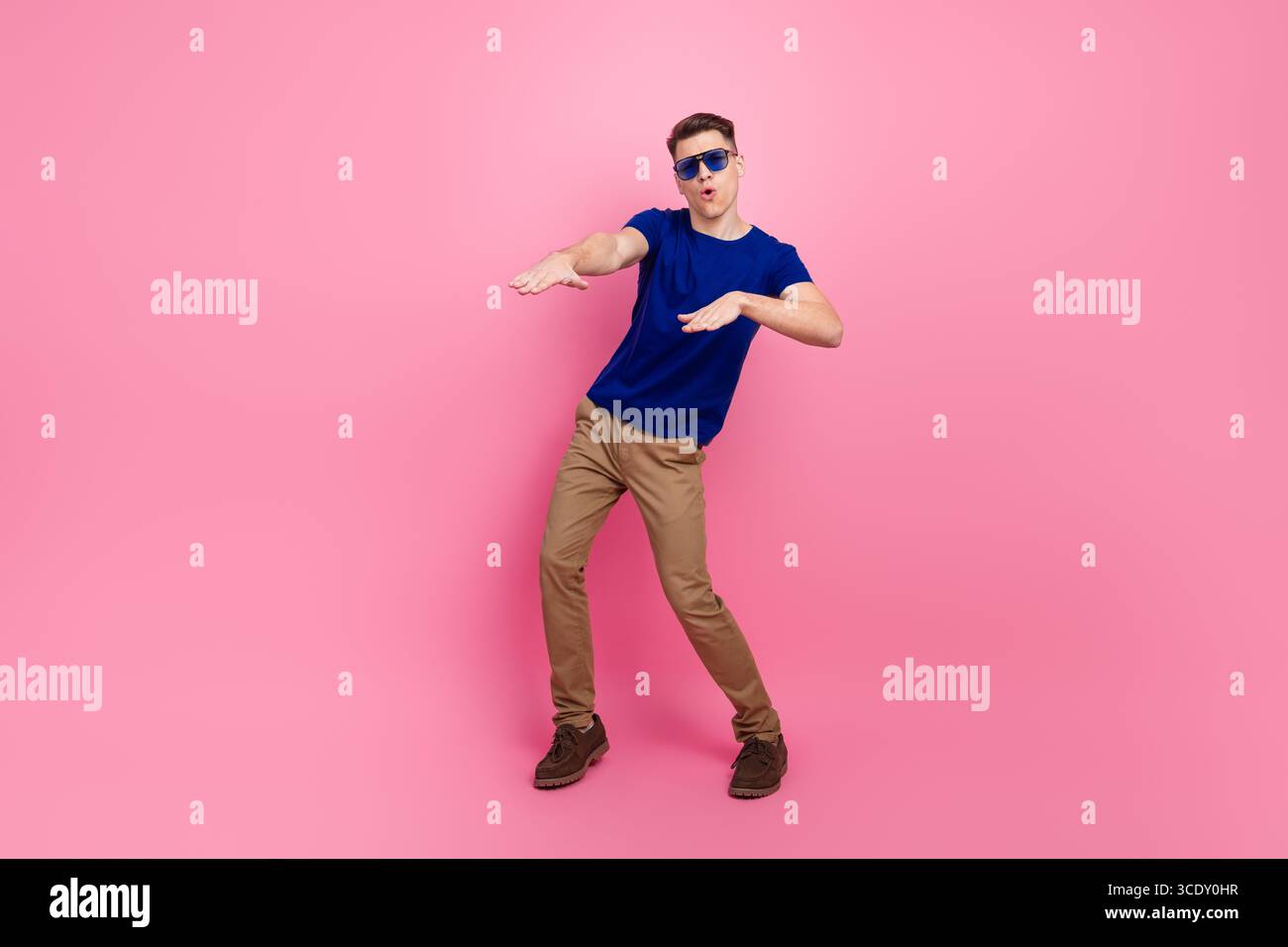 Junger Mann in einem blauen Hemd, der eine verspielte Pose vor einem leuchtend rosa Hintergrund zeigt und Freude und lässigen Stil ausdrückt Stockfoto