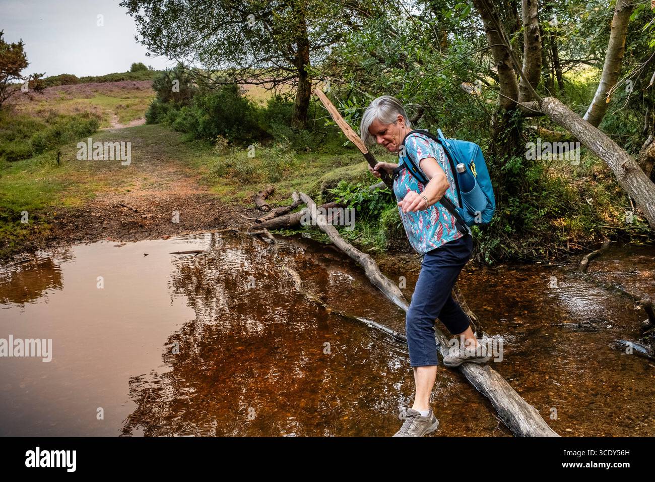 Trail Walker und eine aktive Frau aus den 60er Jahren, die einen Bach ausbalanciert und überwindet und von einer Holzbrücke über das Wasser im New Forest UK springt Stockfoto
