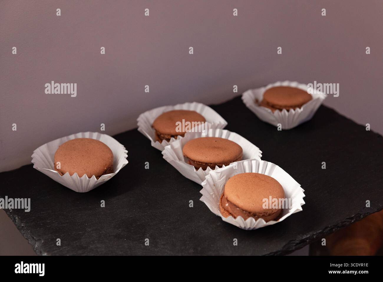 Eine elegante Schoko-Macarons auf dunkler Schieferoberfläche, die jeweils in einem weißen Papierbecher untergebracht sind. Perfekt für Dessertpräsentationen und Kuhgabe Stockfoto