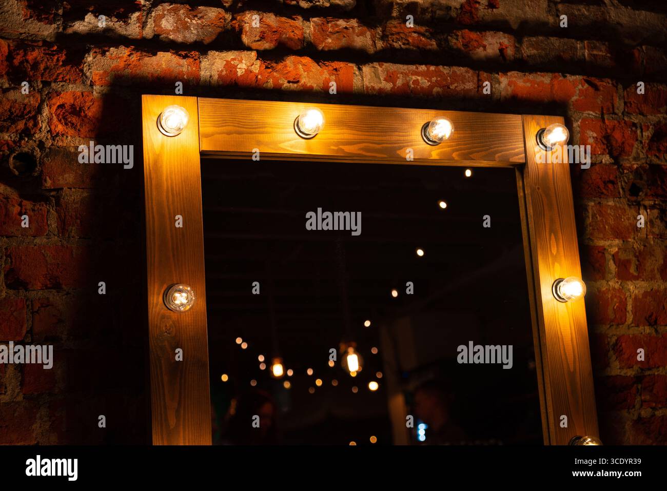 Ein hölzerner Kosmetikspiegel mit warmen, leuchtenden Lichtern, die an einer rustikalen Ziegelwand angebracht sind. Sanfte Umgebungsbeleuchtung sorgt für eine gemütliche und einladende Atmosphäre. Stockfoto