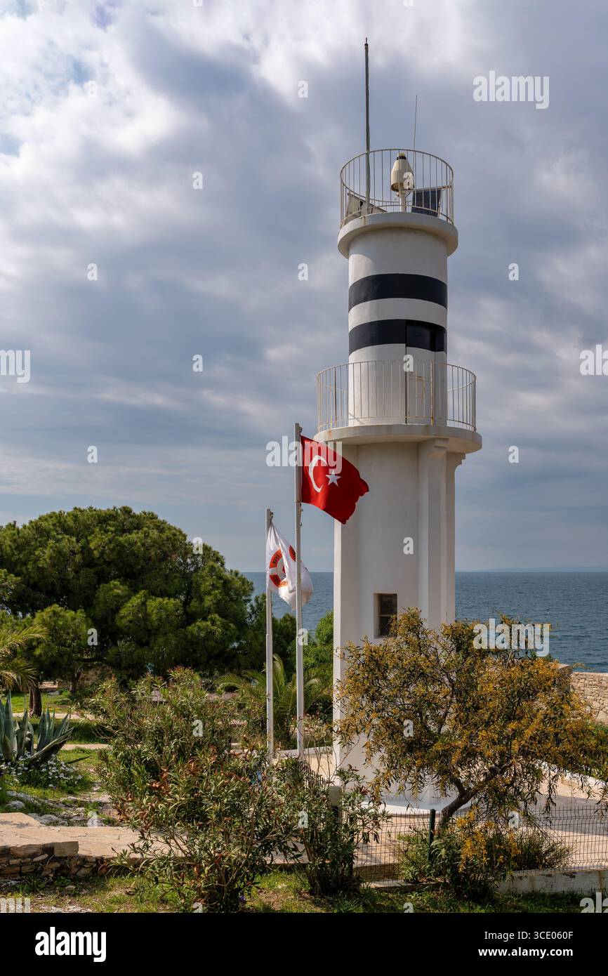Guvercinada Leuchtturm mit türkischer Flagge auf Pigeon Island, Kusadasi, Türkei. Weiß-schwarz gestreifter Leuchtturm an der Ägäis unter bewölktem Himmel Stockfoto