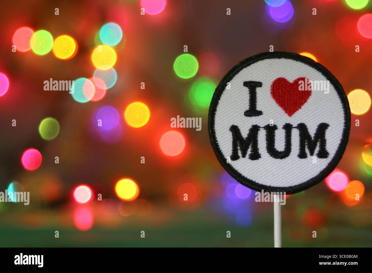 Aufgestickter Patch „I love Mum“ mit rotem Herz auf dem Hintergrund von Regenbogenbokeh-Lichtern, der Platz für Text lässt. Festlicher Muttertag oder Feiertagskonzept. Stockfoto