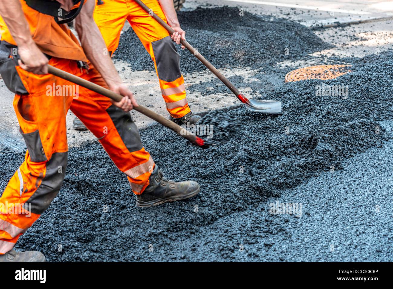 Straßenbau, Straßenbauarbeiten, Arbeiter bessern einen Teil einer innerstädtischen Straße mit Asphalt aus, der heiße Asphalt wird von Hand um einen Kanaldeckel verteilt, anschließend mit einer Straßenwalze verdichtet und geplättet, NRW, Deutschland, Straßenbau *** Straßenbau, Straßenbauarbeiten, Arbeiter reparieren einen Teil einer innerstädtischen Straße mit Asphalt, der heisse Asphalt wird von Hand um einen Mannlochdeckel verteilt, dann mit einer Straßenwalze verdichten und abflacht, NRW, Deutschland, Straßenbau, Straßenbau Stockfoto