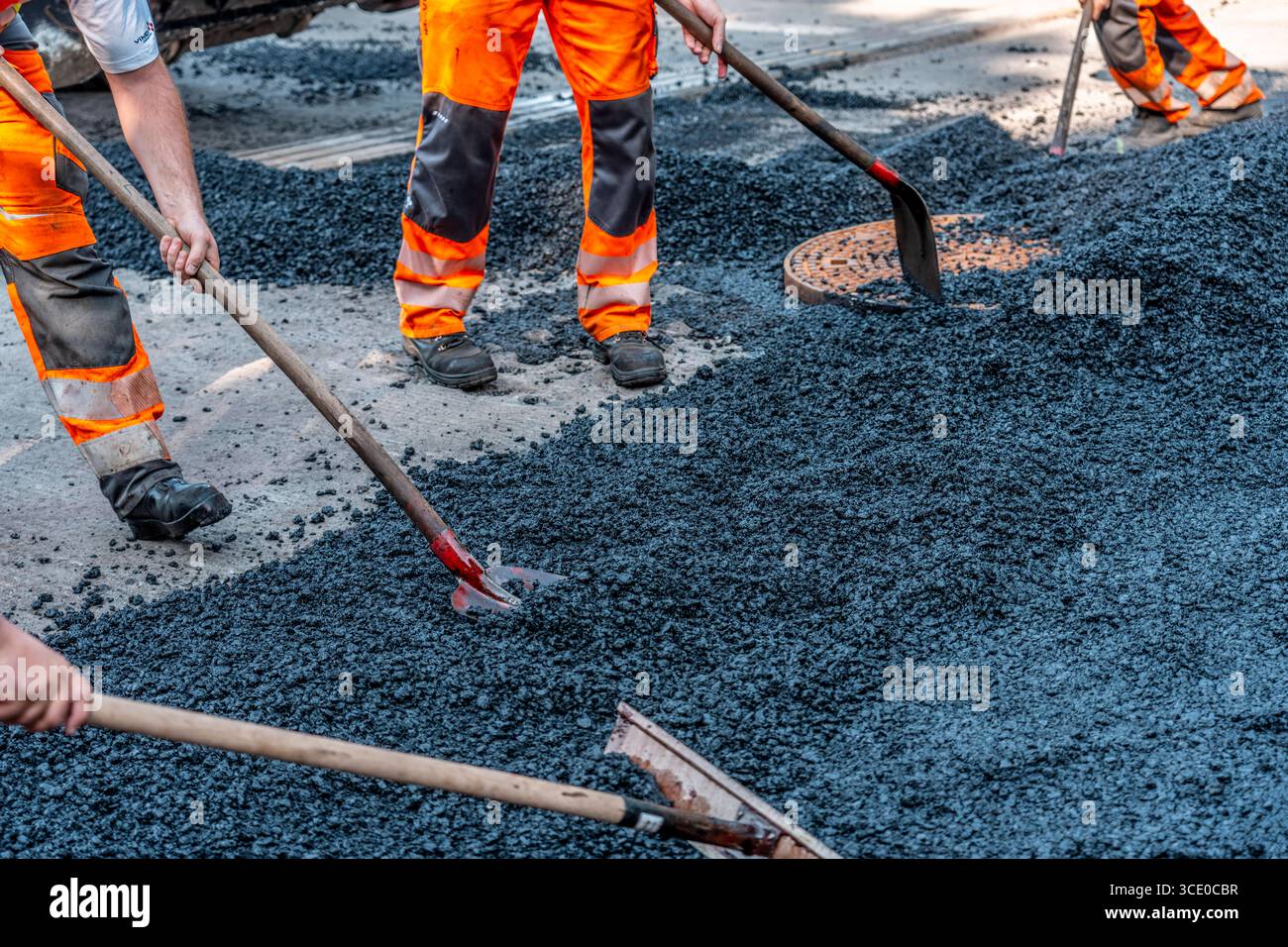 Straßenbau, Straßenbauarbeiten, Arbeiter bessern einen Teil einer innerstädtischen Straße mit Asphalt aus, der heiße Asphalt wird von Hand um einen Kanaldeckel verteilt, anschließend mit einer Straßenwalze verdichtet und geplättet, NRW, Deutschland, Straßenbau *** Straßenbau, Straßenbauarbeiten, Arbeiter reparieren einen Teil einer innerstädtischen Straße mit Asphalt, der heisse Asphalt wird von Hand um einen Mannlochdeckel verteilt, dann mit einer Straßenwalze verdichten und abflacht, NRW, Deutschland, Straßenbau, Straßenbau Stockfoto