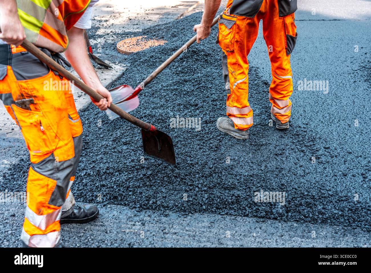 Straßenbau, Straßenbauarbeiten, Arbeiter bessern einen Teil einer innerstädtischen Straße mit Asphalt aus, der heiße Asphalt wird von Hand um einen Kanaldeckel verteilt, anschließend mit einer Straßenwalze verdichtet und geplättet, NRW, Deutschland, Straßenbau *** Straßenbau, Straßenbauarbeiten, Arbeiter reparieren einen Teil einer innerstädtischen Straße mit Asphalt, der heisse Asphalt wird von Hand um einen Mannlochdeckel verteilt, dann mit einer Straßenwalze verdichten und abflacht, NRW, Deutschland, Straßenbau, Straßenbau Stockfoto