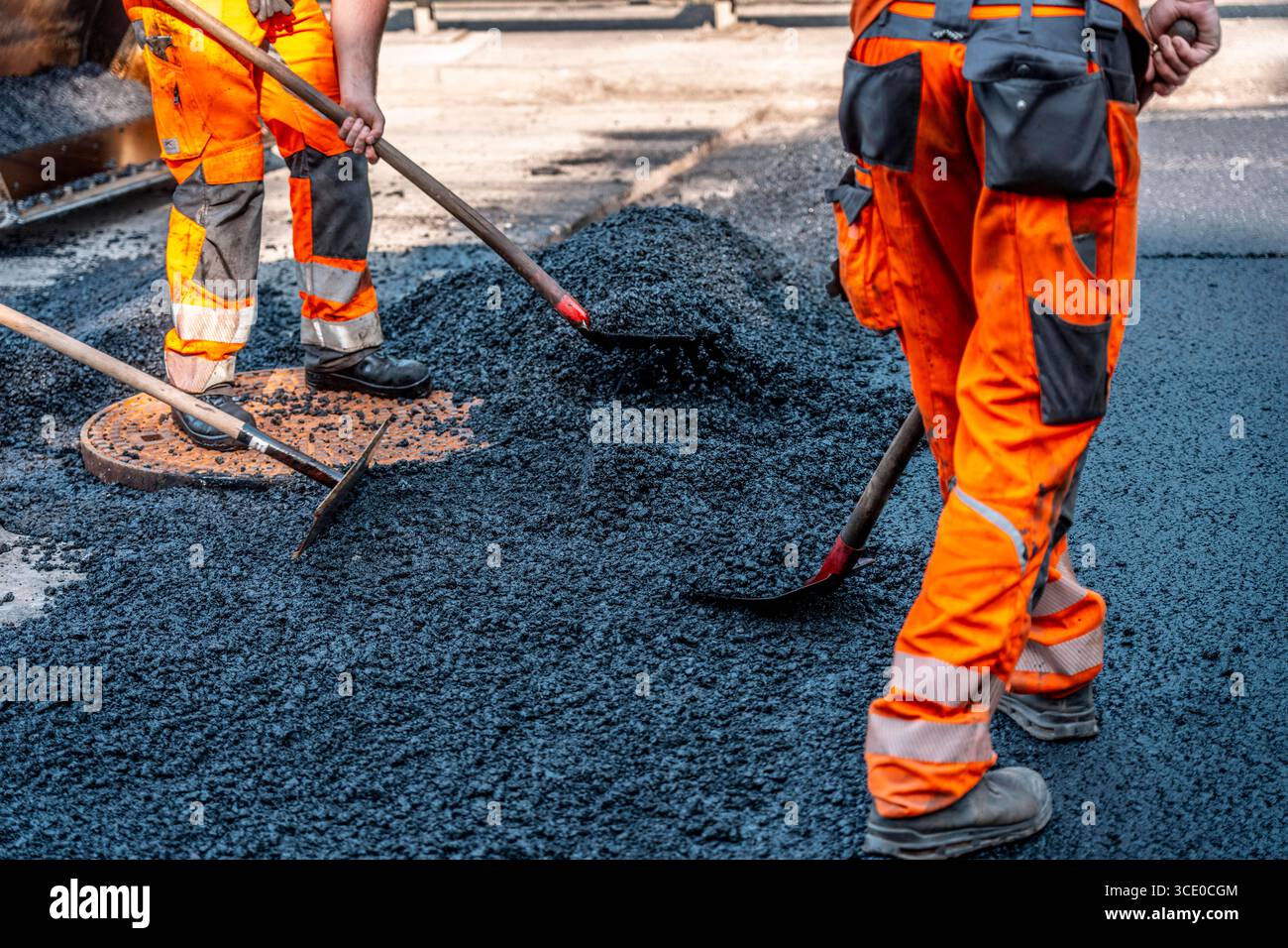 Straßenbau, Straßenbauarbeiten, Arbeiter bessern einen Teil einer innerstädtischen Straße mit Asphalt aus, der heiße Asphalt wird von Hand um einen Kanaldeckel verteilt, anschließend mit einer Straßenwalze verdichtet und geplättet, NRW, Deutschland, Straßenbau *** Straßenbau, Straßenbauarbeiten, Arbeiter reparieren einen Teil einer innerstädtischen Straße mit Asphalt, der heisse Asphalt wird von Hand um einen Mannlochdeckel verteilt, dann mit einer Straßenwalze verdichten und abflacht, NRW, Deutschland, Straßenbau, Straßenbau Stockfoto