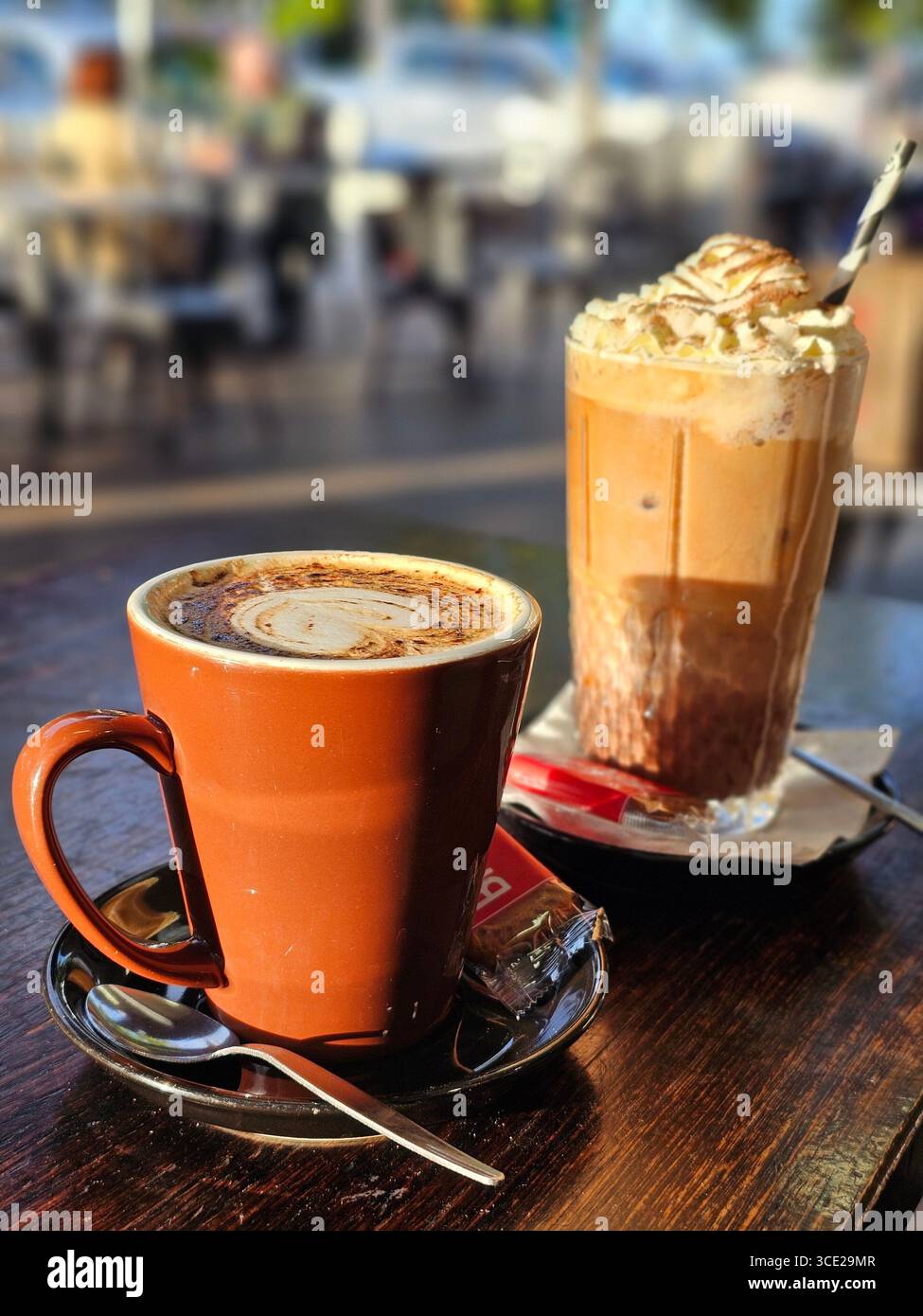 Ein frisch gebrühter Cappuccino in einer Tasse mit Untertasse, Teelöffel und Zucker und sitzt neben einem hohen Glas Eiskaffee mit Schlagsahne. Stockfoto