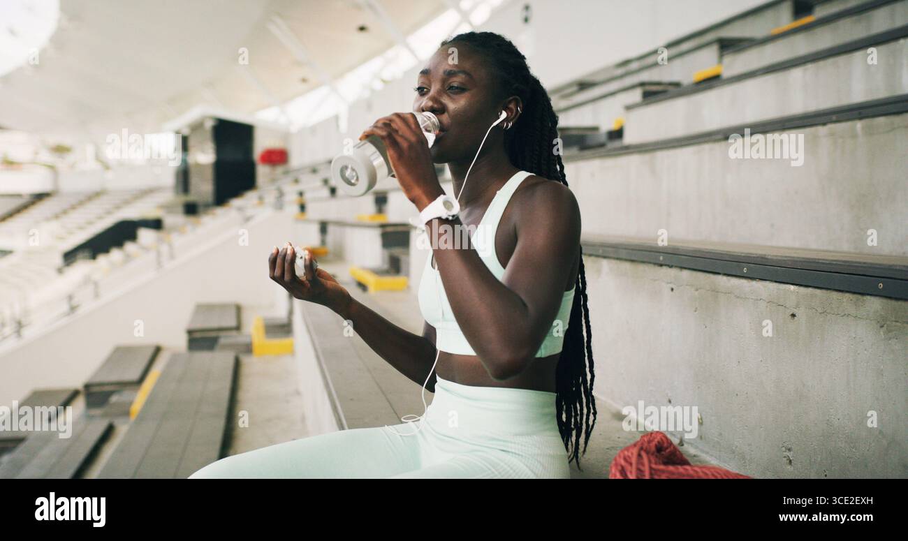 Stadion, schwarze Frau und trinken Sie mit einer Wasserflasche für Fitness, Trinkwasser oder Laufpause. Outdoor, Sportler Person und Gesundheit mit Ohrhörern für Stockfoto