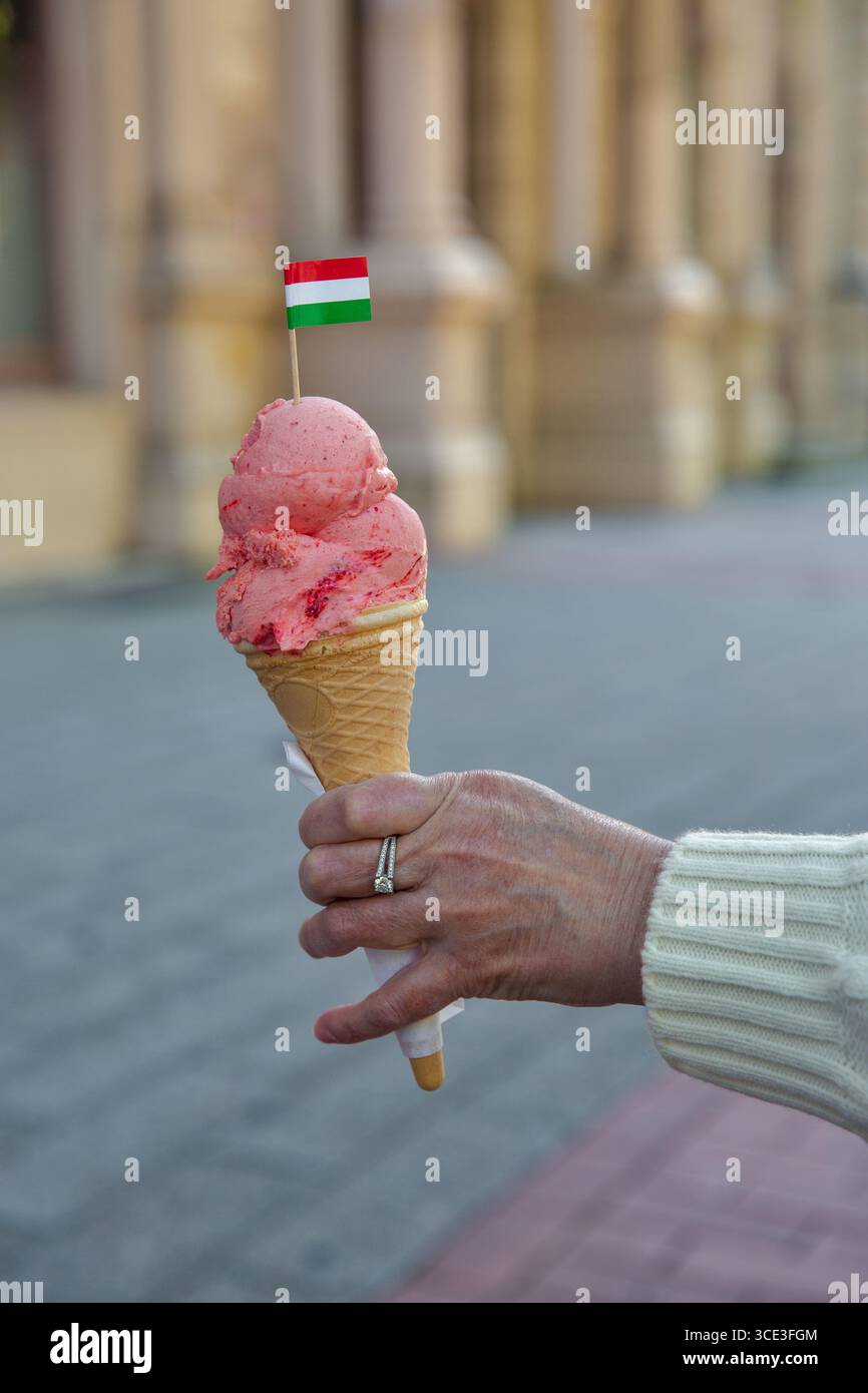 Hand mit einem Waffelkegel mit zwei Kugeln Erdbeereis, gekrönt mit einer kleinen ungarischen Flagge, vor einem verschwommenen Hintergrund der Stadtstraße. Stockfoto