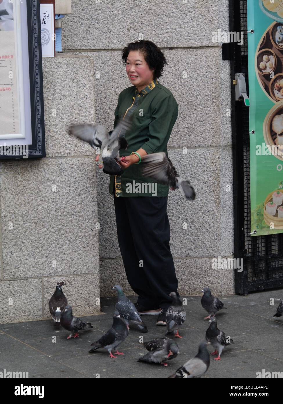 08/2025 — Central London, Vereinigtes Königreich — eine ostasiatische Frau füttert Tauben vor einem chinesischen Restaurant in Central London. In der Hauptstadt treffen sich urbane Tiere und Menschen in einer geschäftigen Straßenszene Stockfoto