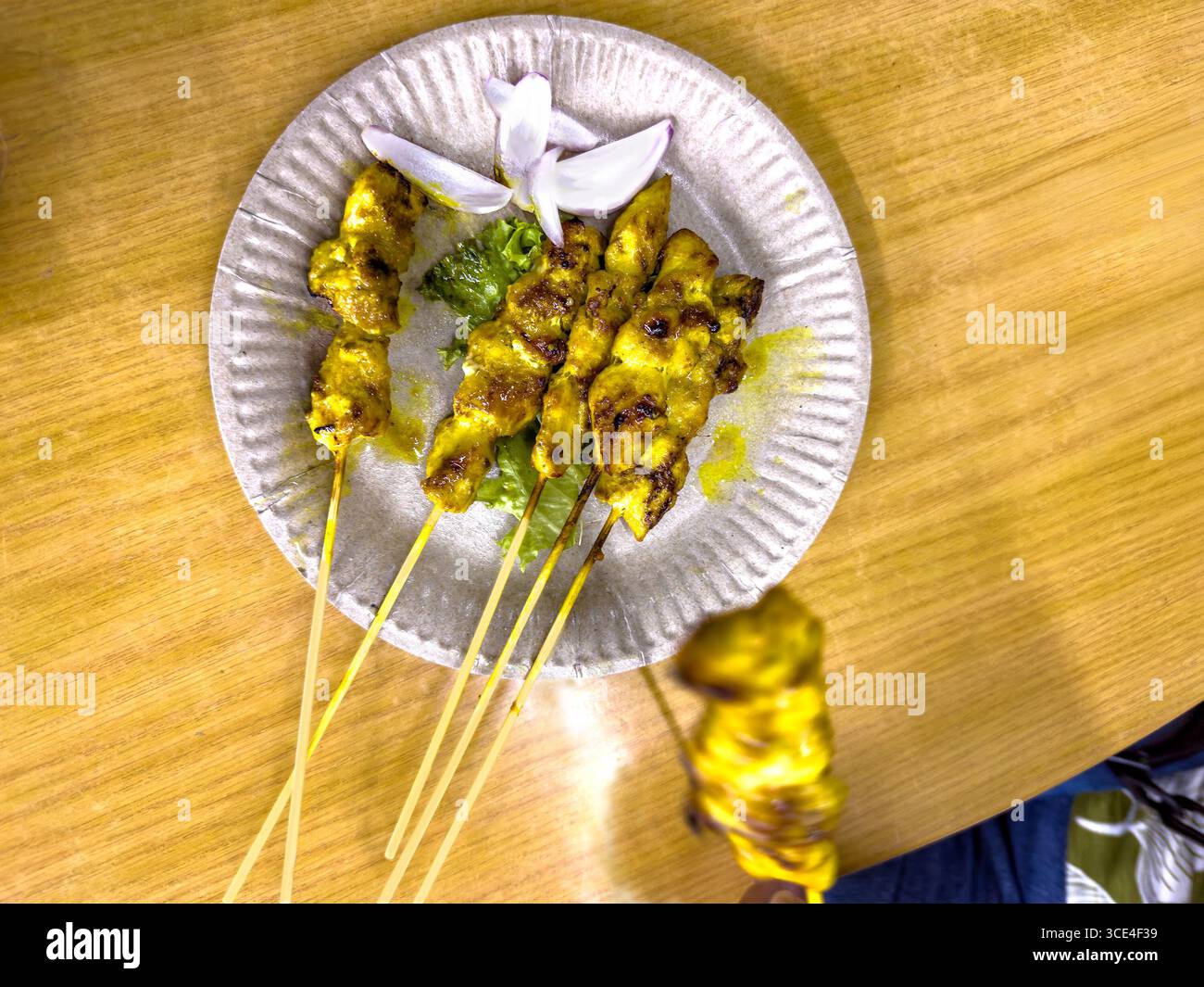Blick von oben auf Satay-Spieße, garniert mit Blumen und Koriander, serviert auf einem Papierteller über einem Holztisch malaysia Stockfoto
