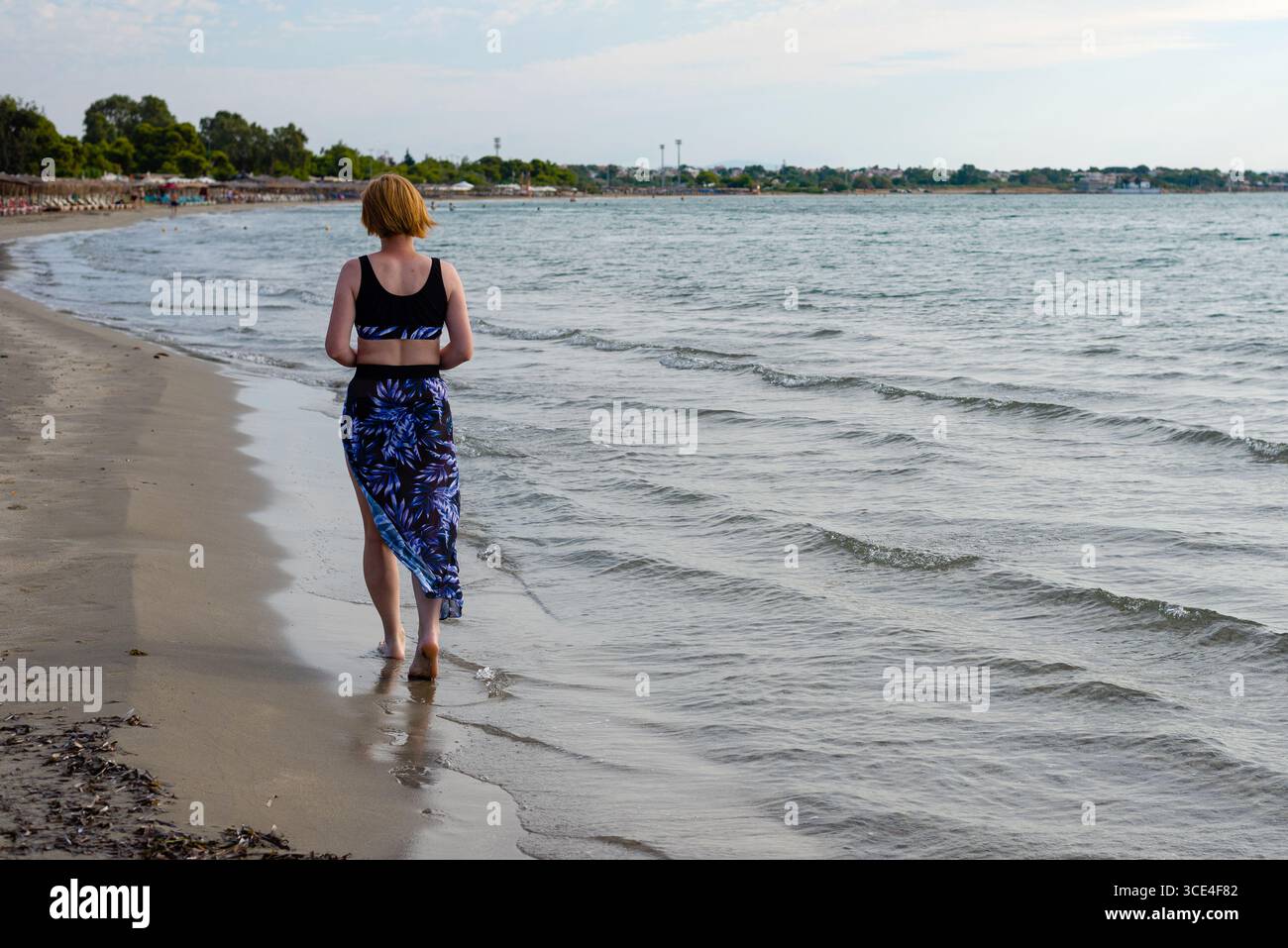 Eine Frau in sportlichem Outfit, die an einem sonnigen Sommertag barfuß an der Küste entlang läuft, mit sanften Wellen, die über den Sand ziehen. Stockfoto