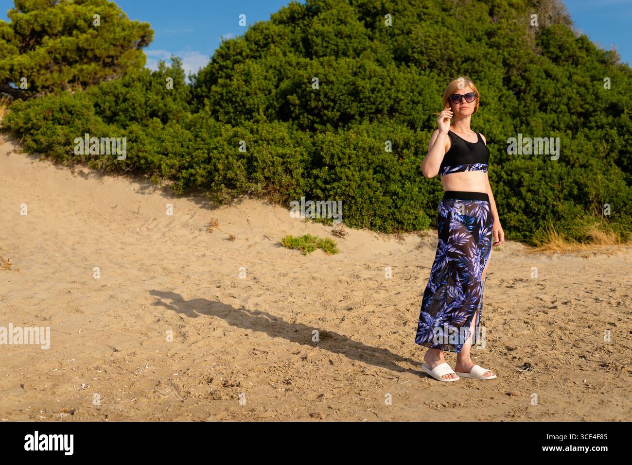 Eine Frau in sportlichem Outfit und Sonnenbrille, die an einem sonnigen Tag auf einem sandigen Hügel mit üppigen grünen Büschen im Hintergrund steht. Stockfoto