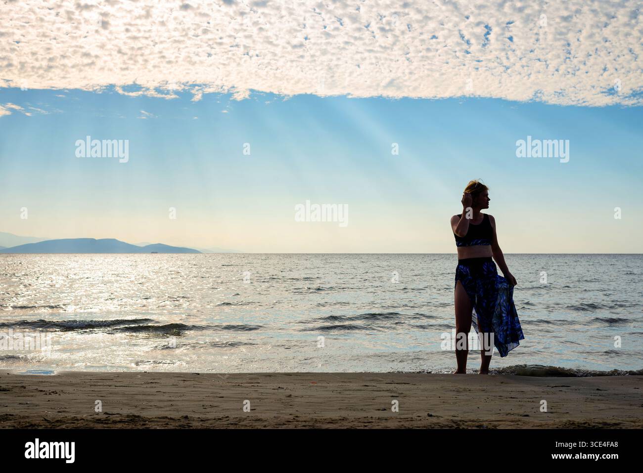 Eine Frau in sportlichem Outfit, die ein blaues Hemd in der Hand hält, am Sandstrand steht und bei sonnigem Wetter auf den Meereshorizont blickt. Stockfoto