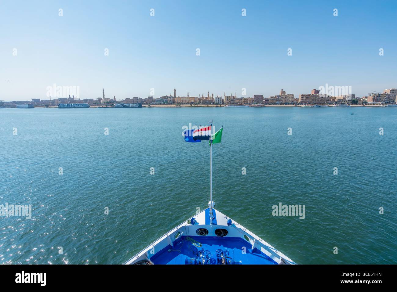 Aus nächster Nähe vom Bug eines Kreuzfahrtschiffes auf dem Nil, mit der ägyptischen und Luxor-Flaggen, die mit der fernen Stadtlandschaft von Luxor un fliegen Stockfoto