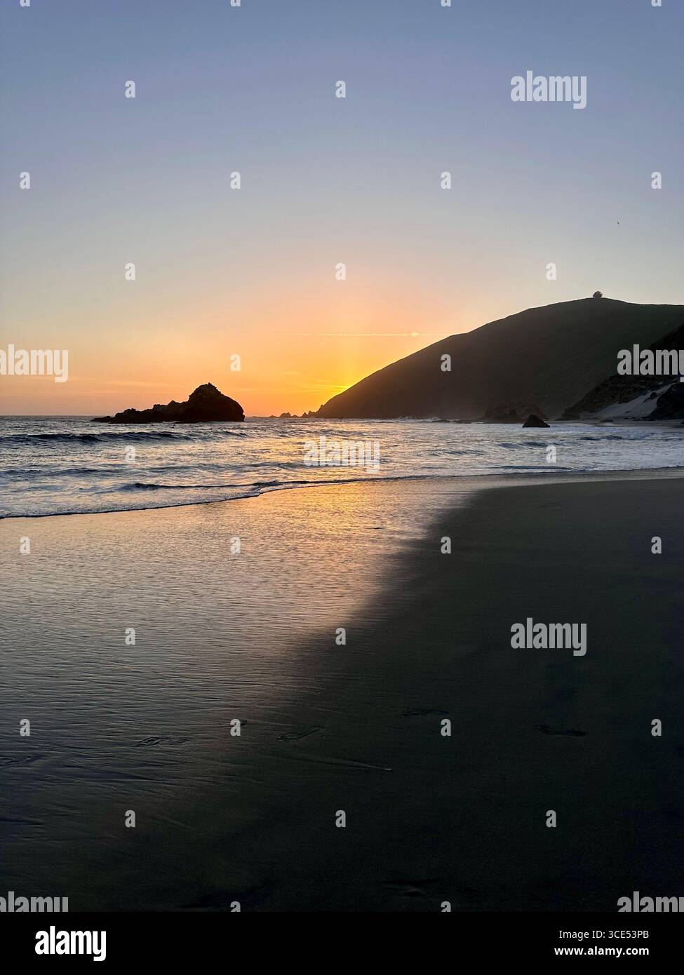 Goldener Sonnenuntergang über dem Pazifischen Ozean mit Wellen, die auf den Sandstrand und zerklüftete Klippen im Hintergrund schweben. - Smartphone-aufgenommenes Stockfoto