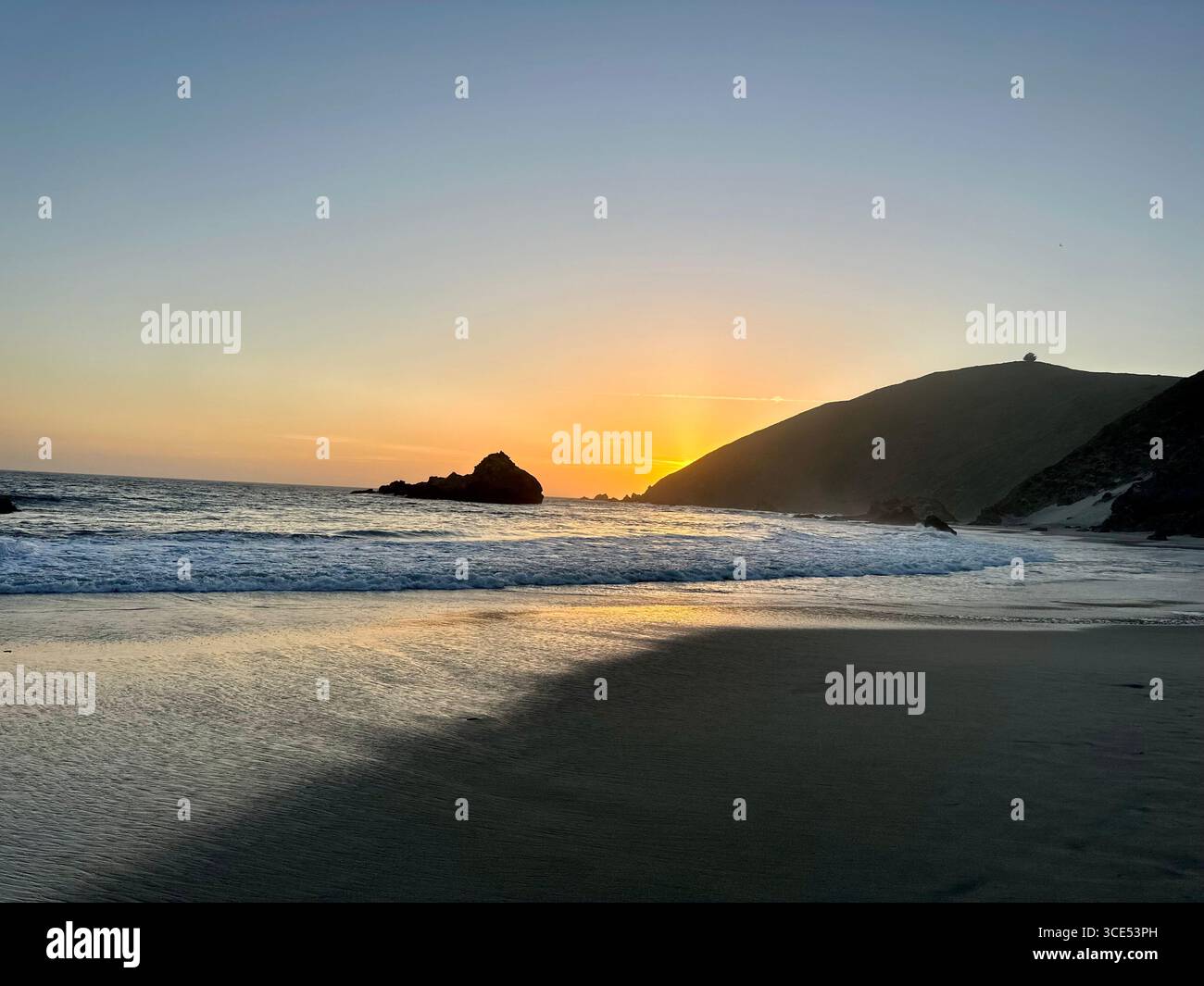Goldener Sonnenuntergang über dem Pazifischen Ozean mit Wellen, die auf den Sandstrand und zerklüftete Klippen im Hintergrund schweben. - Smartphone-aufgenommenes Stockfoto