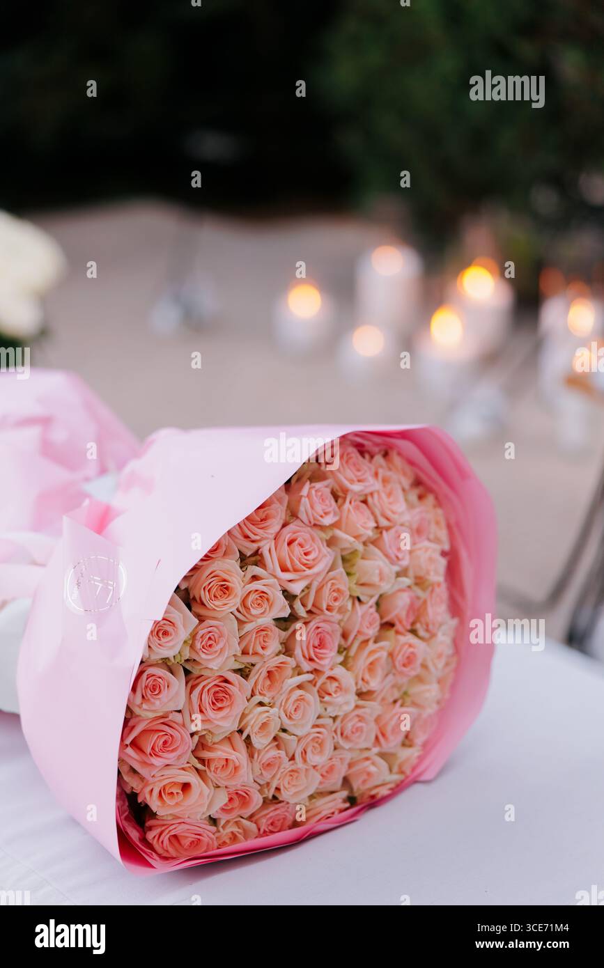 Luxuriöser großer Strauß rosafarbener Rosen, gewickelt in weichem rosa Papier, liegt auf einem Tisch, im Gartenhintergrund mit verschwommenen Glühkerzen. Romantisch und Stockfoto