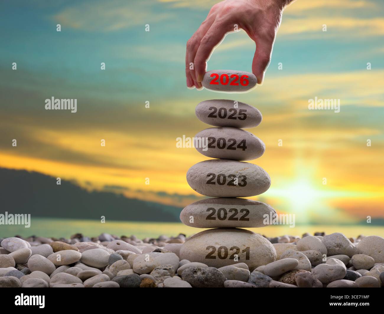 Mit gestapelten Steinen an einem Strand bei Sonnenuntergang, gekennzeichnet mit Jahren von 2021 bis 2026. Eine Hand legt den mit 2026 markierten oberen Stein Stockfoto