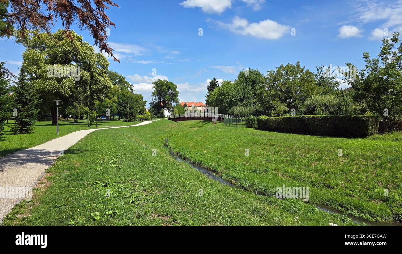 Ein sanfter Bach mit einer kleinen Fußgängerbrücke und gewundenen Parkwegen ist von üppigen grünen Rasenflächen, Reifen Bäumen und blauem Himmel umgeben und lädt zu einem malerischen Spaziergang ein Stockfoto
