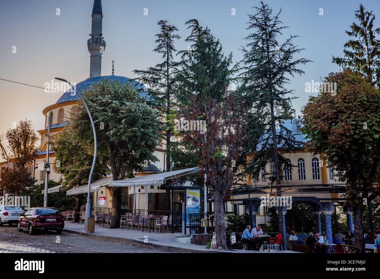 Traditionelle Moschee mit blauer Kuppel und hohem Minarett, umgeben von Bäumen, Geschäften und Menschen in Cafés im Freien in Başiskele, Kocaeli, Türkiye, während su Stockfoto