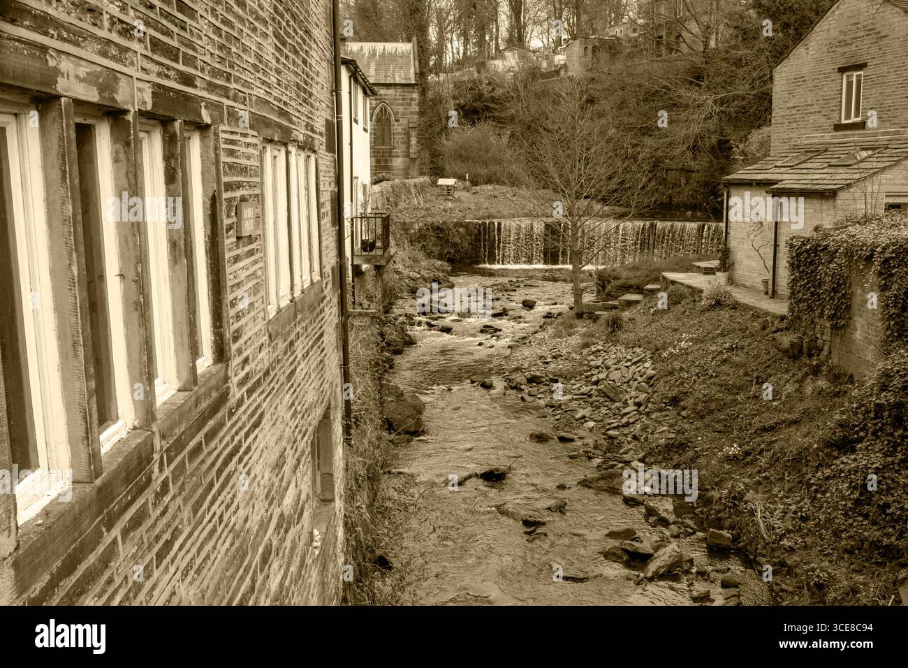 Ein sepiafarbener Blick auf den Luddenden Bach, der zwischen traditionellen Steinbauten fließt, mit einem kleinen Wasserfall im Hintergrund. Stockfoto