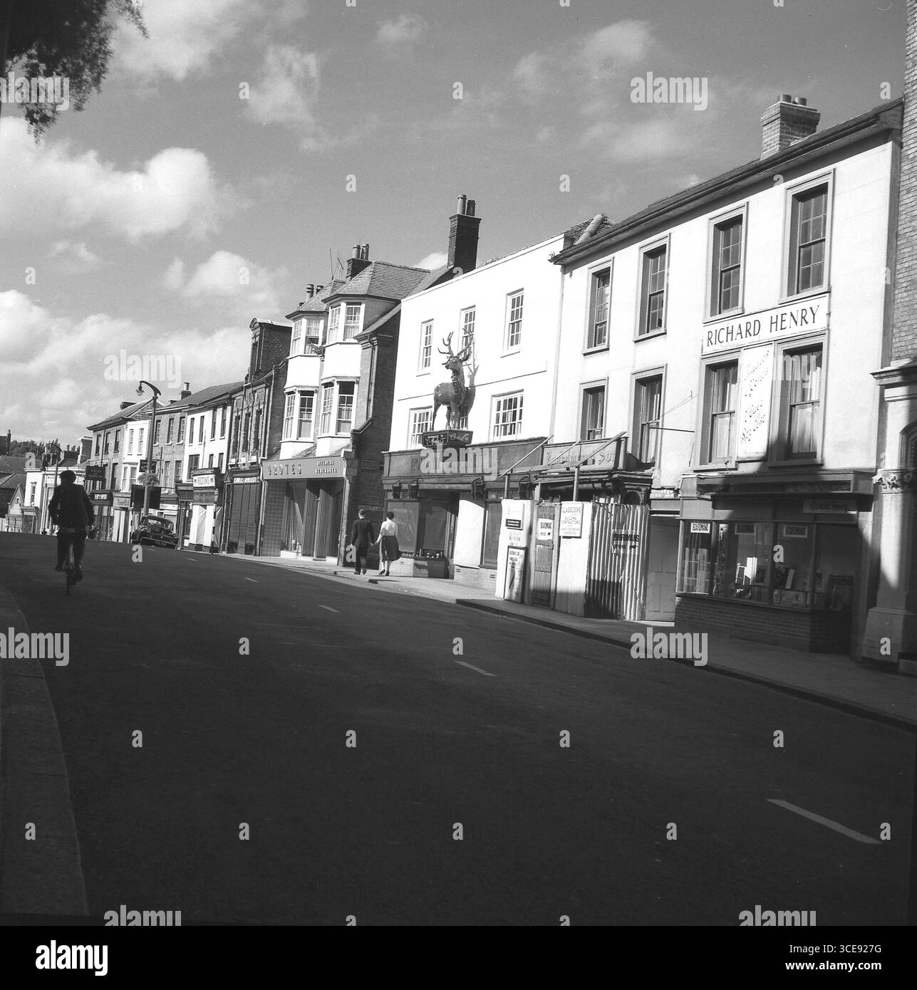 1950er Jahre, historisches Bild der High Street, Bishop's Stortford, England, Großbritannien. Zu den Geschäften gehören Holland und Barrett, Richard Henry Friseure, Grove Builders Merchants & Boardmans. Holland und Barrett waren in dieser Zeit ein Lebensmittelgeschäft. Oberhalb des Eingangs befindet sich das ursprüngliche White-Hart-Emblem des 1. Herts Light Horse-Regiments, das 1862 für die Kaserne des Regiments in Silver Leys hergestellt wurde. Als sich das Regiment 1868 auflöste, holte Major William Holland den Hirsch zurück und ließ ihn später über seinem Laden an der Ecke Barrett Lane montieren. Stockfoto