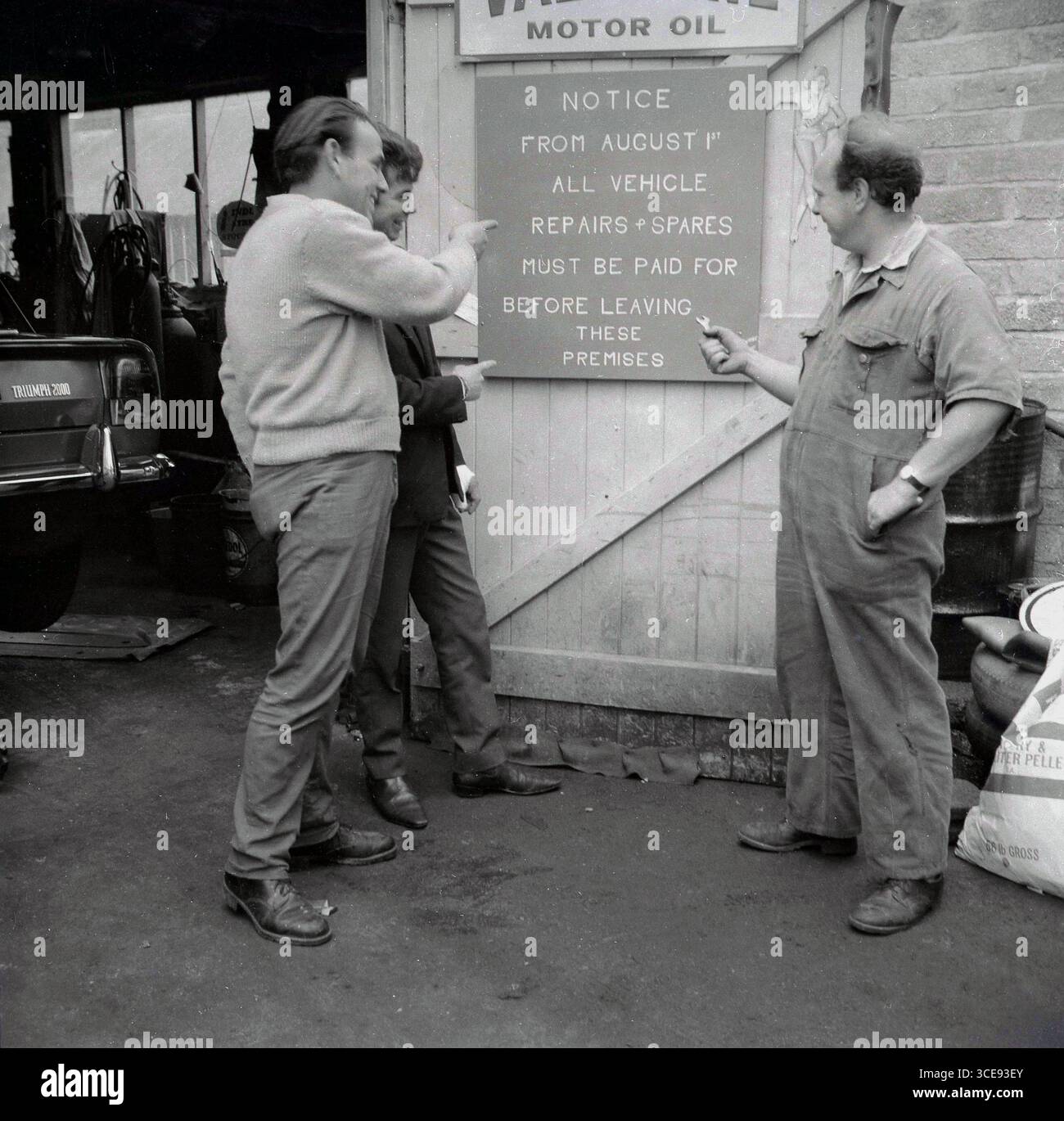 1960er Jahre, historisch, drei Männer, einer ein Mechaniker, vor dem Eingang zu einer kleinen Dorfgarage, lachend und auf einen Hinweis an der hölzernen Garagentür zeigen, der besagt: "Ab dem 1. August müssen alle Fahrzeugreparaturen und Ersatzteile bezahlt werden, bevor sie diese Räumlichkeiten verlassen." Ein Triumph 2000-Auto auf einem Gepäckträger. Stockfoto