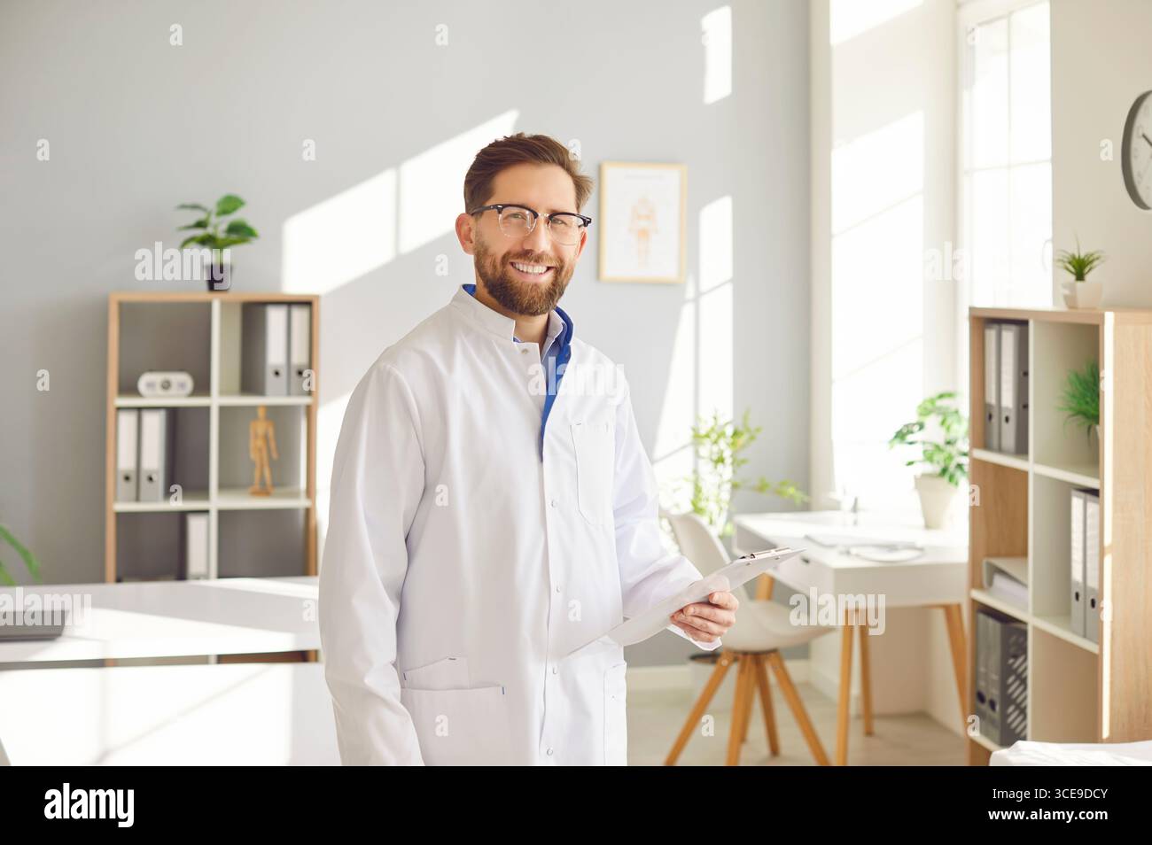 Porträt eines jungen glücklichen männlichen Arztes in weißer medizinischer Uniform, der in seiner Praxis steht. Stockfoto
