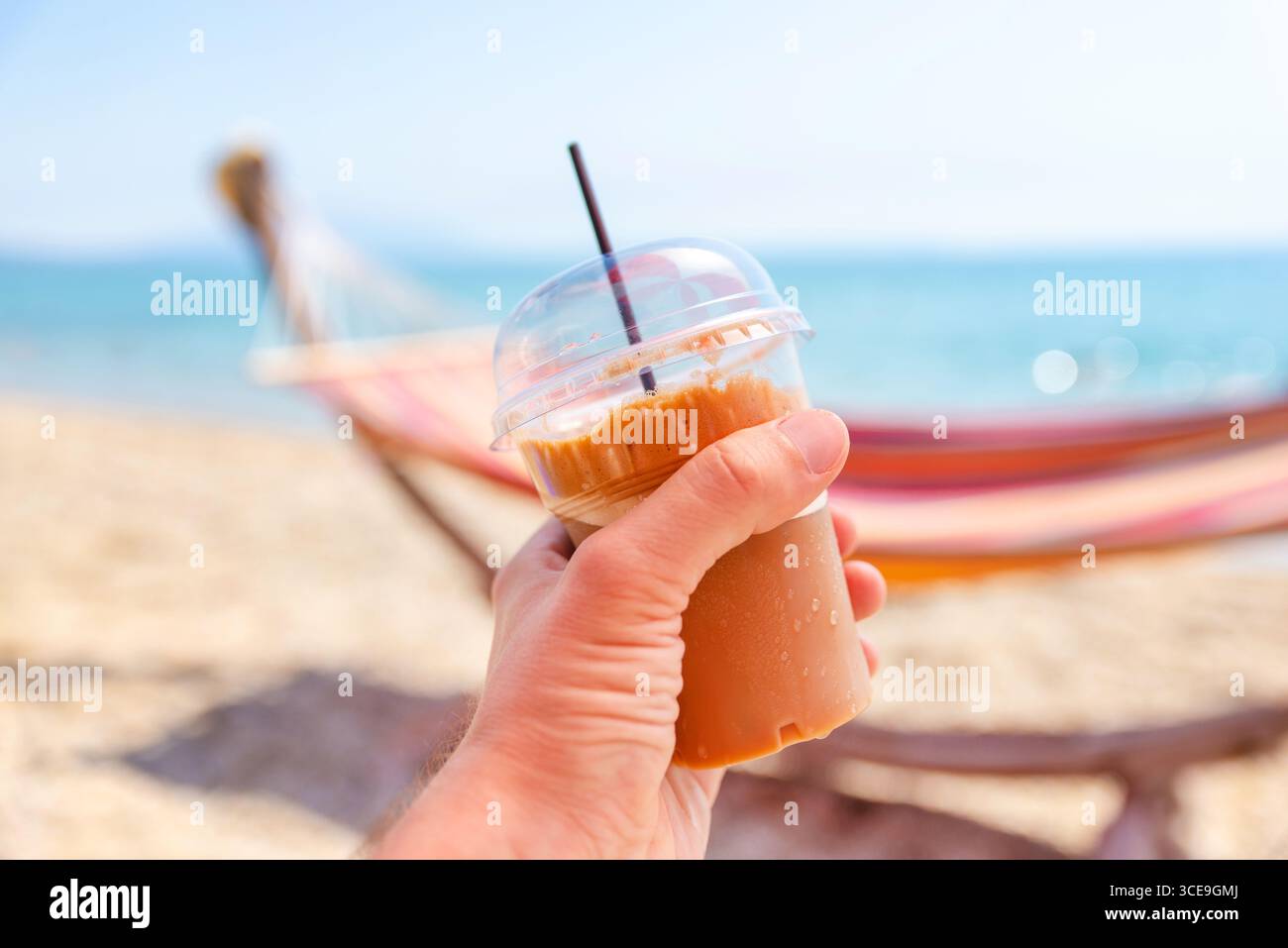 Hand hält Eiskaffee in einer Tasse zum Mitnehmen an einem sonnigen Strand, farbenfrohe Hängematte verschwimmt im Hintergrund Stockfoto