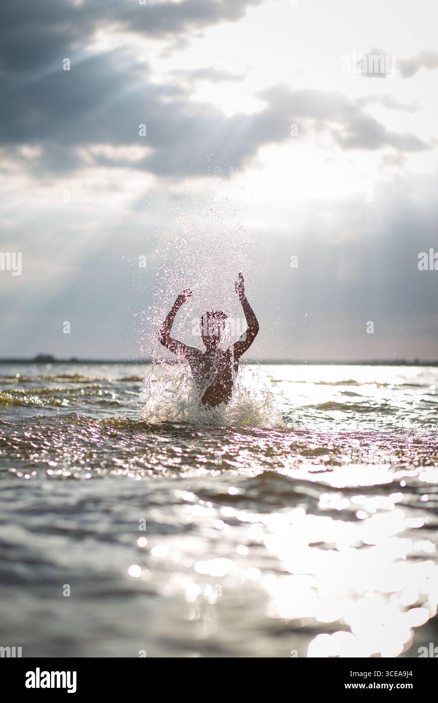Wasser spritzt um eine erhöhte Silhouette, dramatische Sonnenstrahlen durch Wolken, starker Kontrast und glitzernde Sommerenergie. Stockfoto
