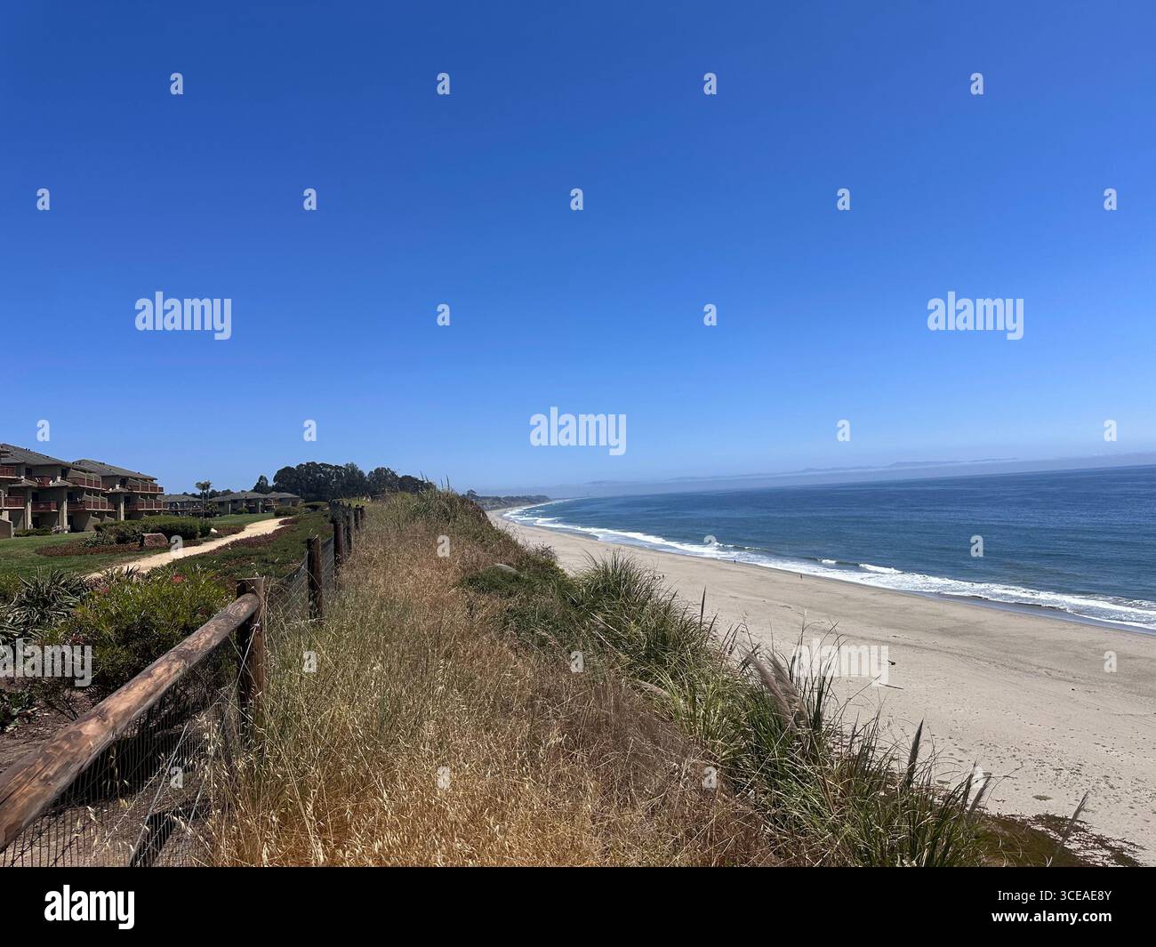 Ein malerischer Blick auf die Küste von einer Klippe mit grasbewachsenen Dünen, Häusern am Meer und einem langen Sandstrand, der auf das ruhige blaue Wasser trifft - Smartphone-aufgenommenes Stockfoto