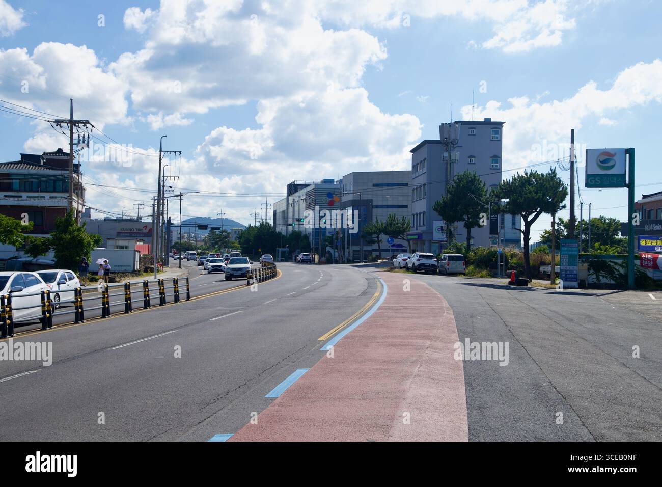 Auf der Nordseite von Jeju führt eine breite, markierte Straße mit Radweg vorbei an Tankstellenbeschilderungen und flachen Unternehmen unter verstreuten Wolken. Stockfoto