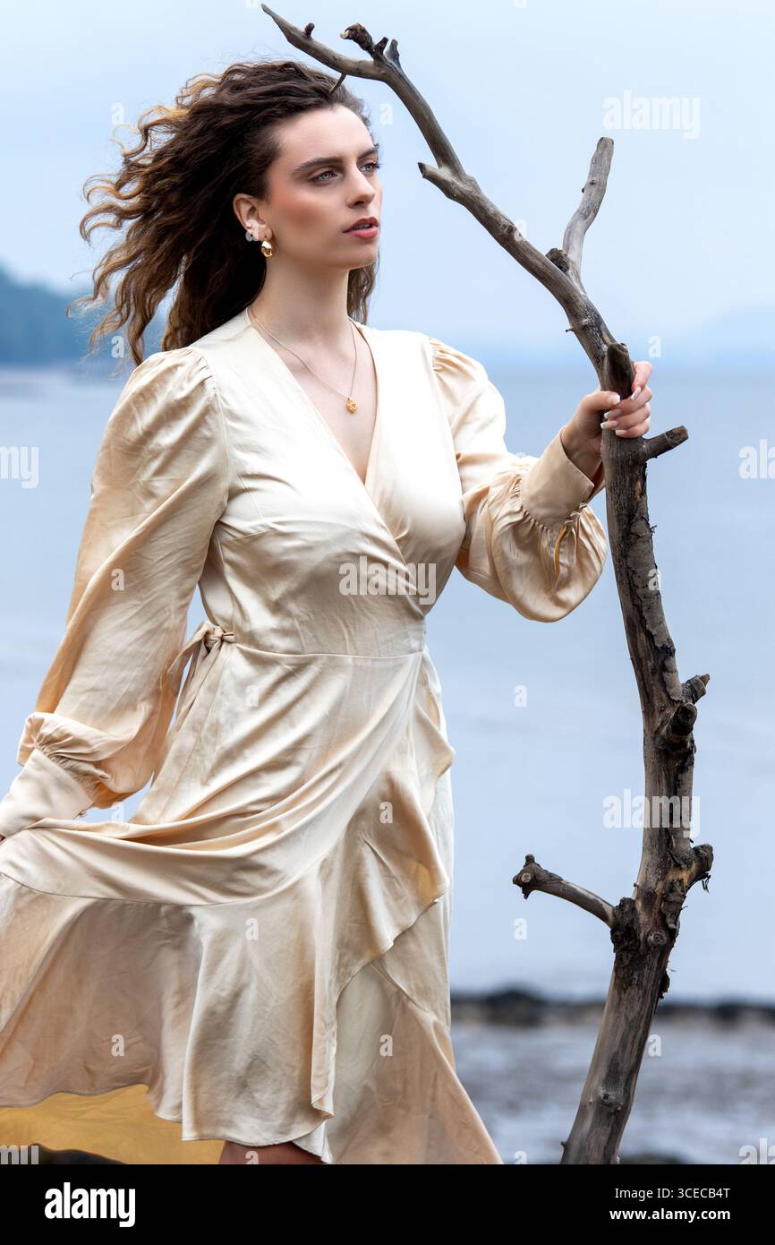 Fantasy-Shooting „Lady of the Lake“ am Wormit Beach, Fife, Schottland, während die Frau in Seidengewändern an den Felsen posiert, die Treibholz in der Hand halten Stockfoto