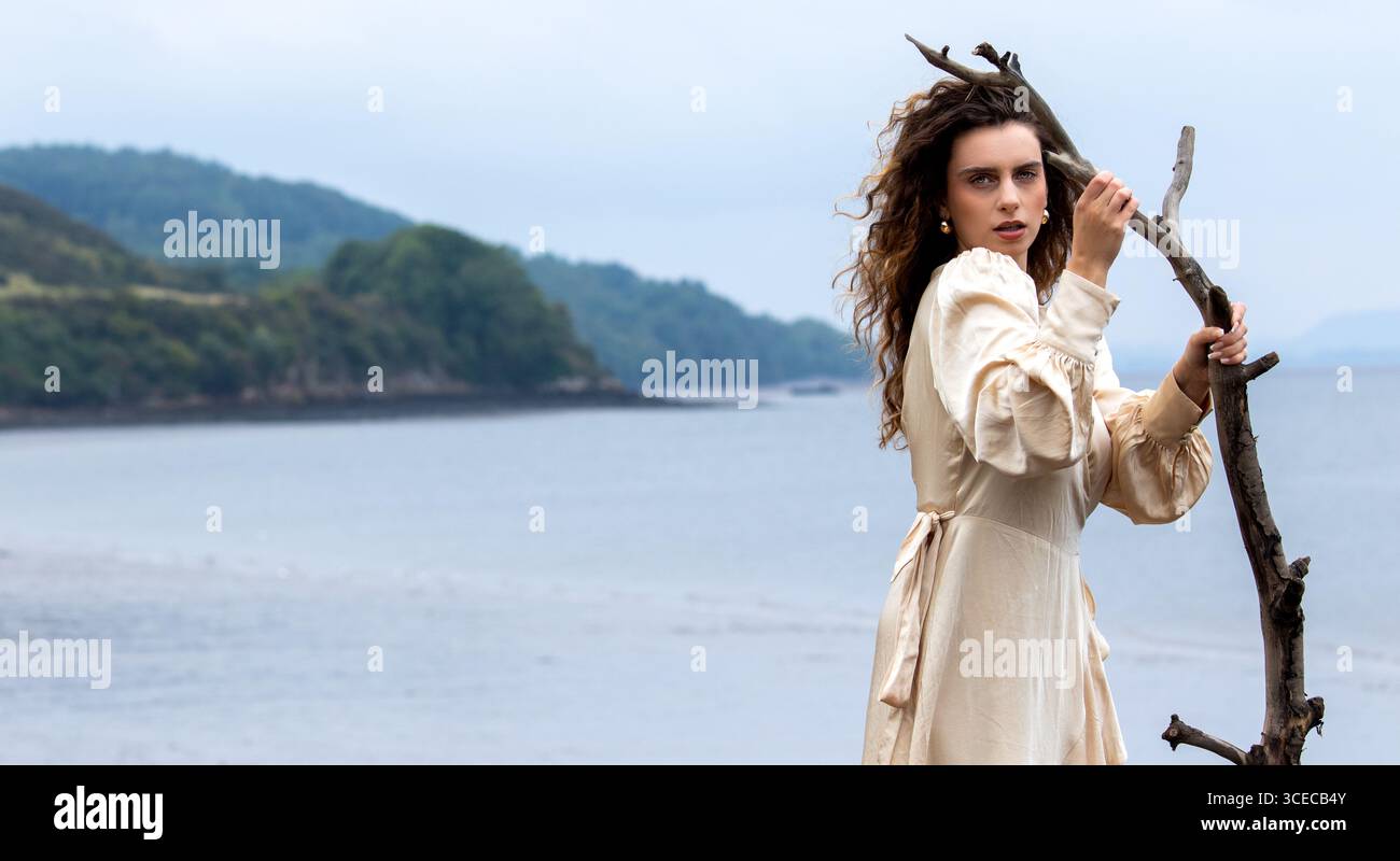 Fantasy-Shooting „Lady of the Lake“ am Wormit Beach, Fife, Schottland, während die Frau in Seidengewändern an den Felsen posiert, die Treibholz in der Hand halten Stockfoto