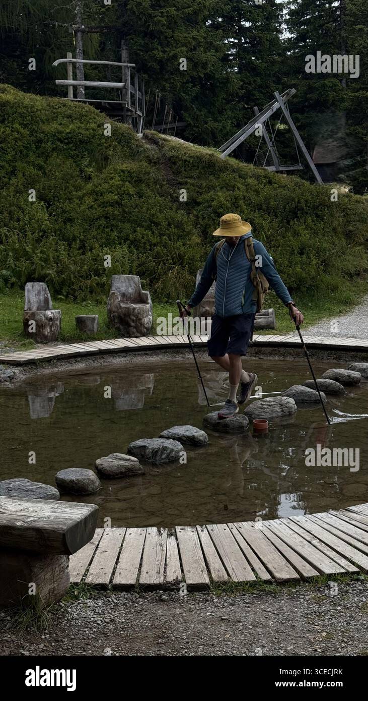 Ein Mann mit Wanderausrüstung überquert vorsichtig eine Reihe von Trittsteinen in einem Waldteich und benutzt Trekkingstöcke für das Gleichgewicht, umgeben von Grün und Grün Stockfoto