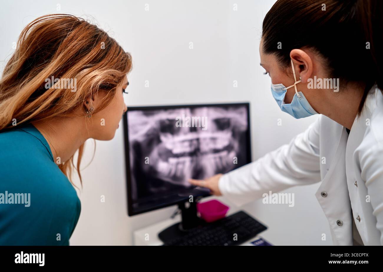 Ein Zahnarzt in einer Zahnklinik zeigt einem Patienten ein Röntgenbild auf einem Computerbildschirm und erklärt Details, während beide sich auf das Bild konzentrieren. Der Patient hört ein Stockfoto