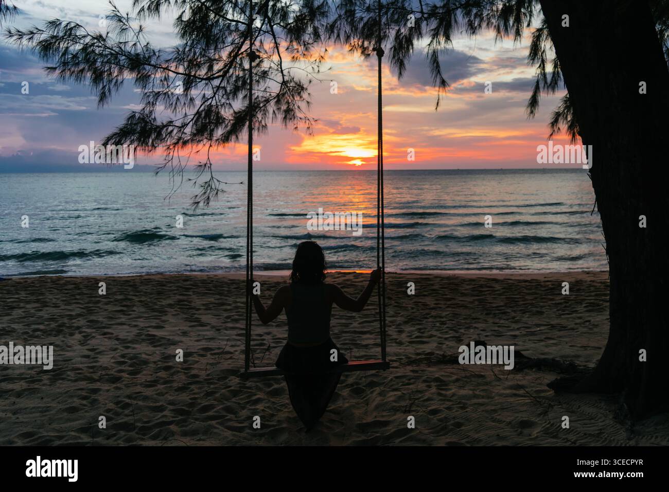 Eine Frau sitzt auf einer Schaukel mit Blick auf den ruhigen Ozean bei Sonnenuntergang auf Koh Rong Island, Kambodscha. Die ruhige Szene fängt den lebendigen Himmel und die Ruhe ein Stockfoto