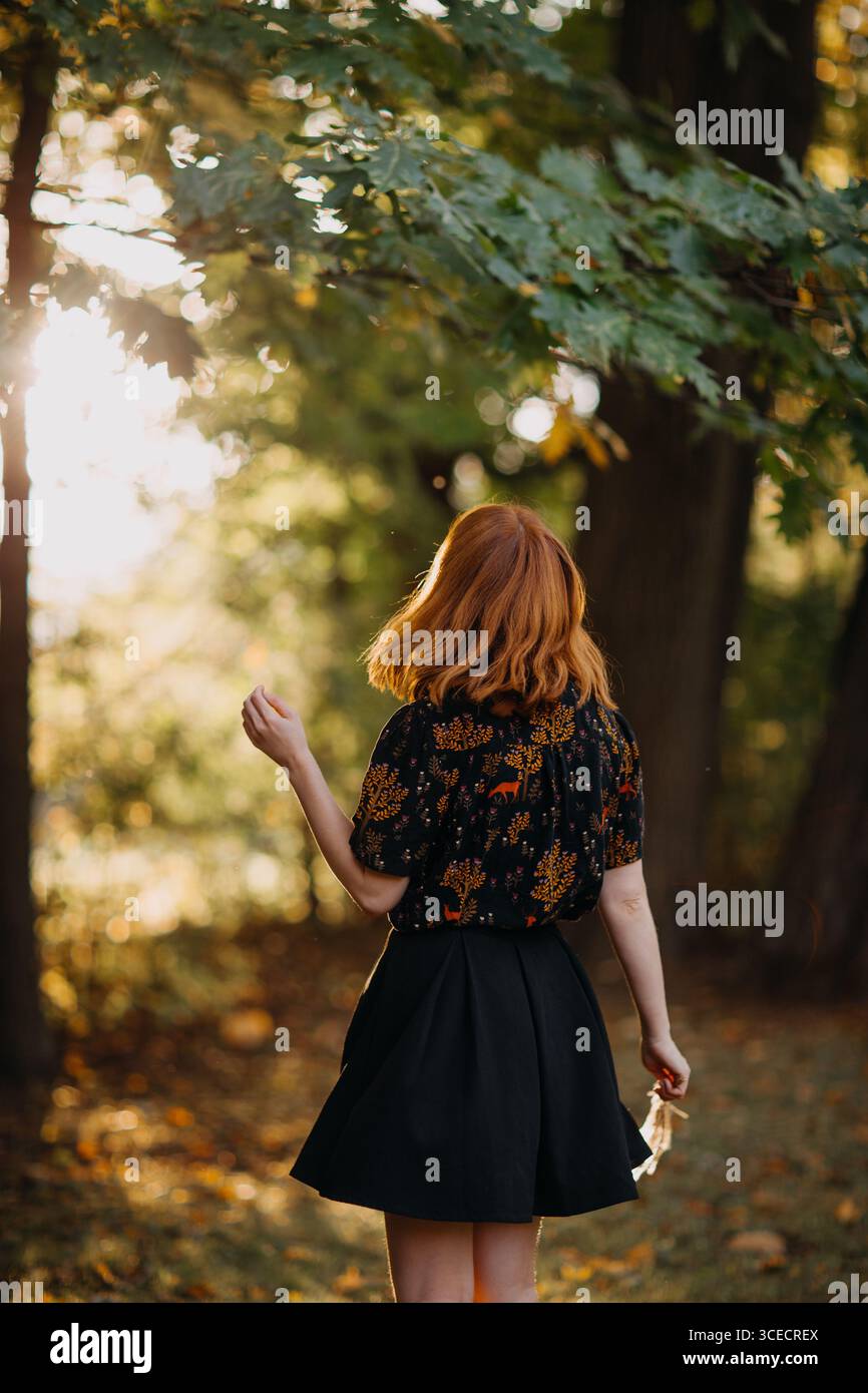 Eine Frau mit rotem Haar genießt das herbstliche Sonnenlicht, das durch Bäume in einem ruhigen Wald von Quebec filtert. Die Szene fängt das Wesen des Falls in Kanada ein. Stockfoto