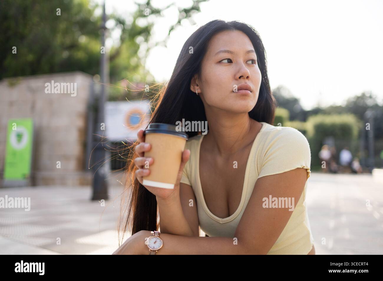 Junge Chinesin, die eine Tasse Kaffee hält, sitzt draußen in einer Stadt. Die Sonne hebt ihr langes Haar hervor, während sie eine entspannte Sommerstimmung genießt Stockfoto