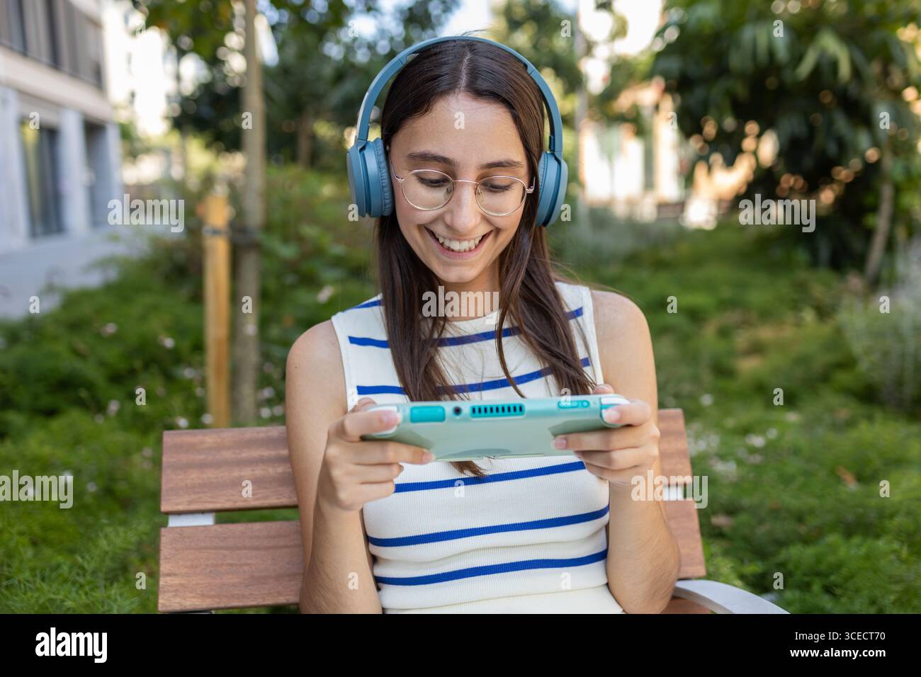 Eine junge Frau entspannt sich auf einer Bank im Stadtpark, trägt Kopfhörer und spielt eine Spielkonsole. Sie liebt das urbane Outdoor-Leben und genießt Ruhe Stockfoto