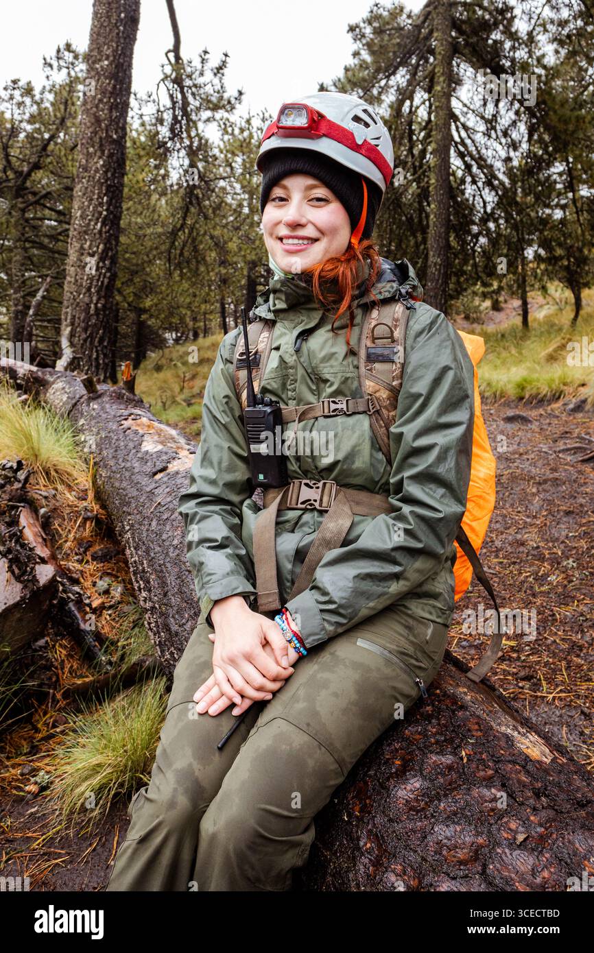 Eine Wanderer sitzt auf einem Baumstamm auf einem üppigen, regnerischen Waldweg und lächelt in wasserdichter Kleidung. Die nebelige Atmosphäre unterstreicht das abenteuerliche Stockfoto
