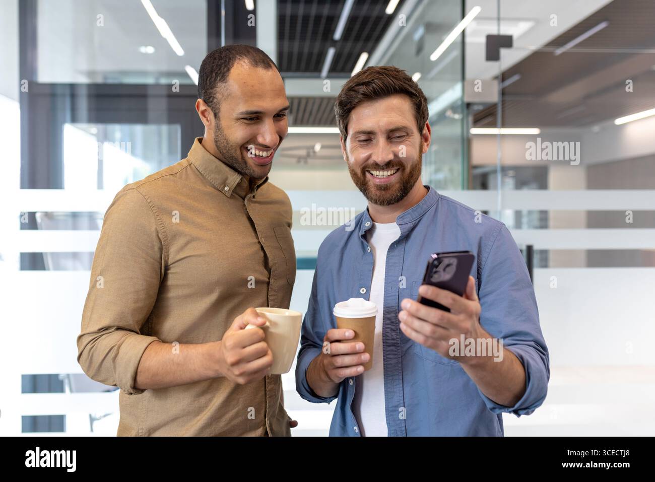 Zwei Kollegen verbringen einen zwanglosen Moment beim Kaffeegenuss, während sie gemeinsam ein Smartphone in einer modernen Büroumgebung betrachten. Dieses Bild zeigt Teamarbeit, Technologie und freundliche Interaktionen Stockfoto