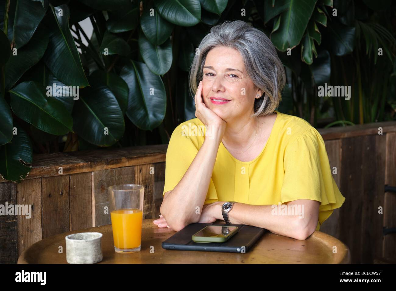 Eine reife Frau mit grauem Haar, die eine gelbe Bluse trägt, sitzt an einem Holztisch in gemütlicher Atmosphäre mit einem Smartphone und einem Glas Saft und strahlt Ruhe aus Stockfoto